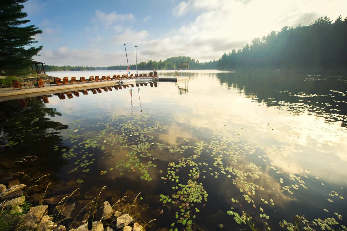 Adirondack and lounge chairs sitting on a wooden dock facing a calm lake during a sunny summer day. Sailing boats are tied to the dock. A stock photo.